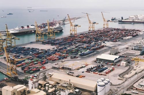 Expansive aerial view of a bustling container port with large cranes and cargo ships under a cloudy sky.